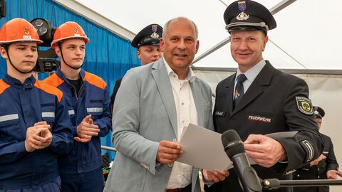 Ein Mann übergibt weißen Mappe an Feuerwehrmann. Im Hintergrund stehen weitere Feuerwehrmänner. 