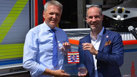 Zwei Männer stehen vor Feuerwehrauto und lachen in Kamera.