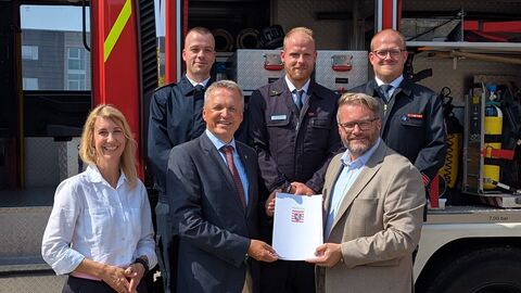Staatssekretär Martin Rößler steht mit weitere Personen vor einem Feuerwehrfahrzeug und hält einen Zuwendungsbescheid in den Händen