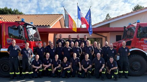 Gruppenfoto von knapp 30 uniformierten Feuerwehrkräften. Im Hintergrund 2 Feuerwehrfahrzeuge sowie die deutsche, die französische und die europäische Flagge.