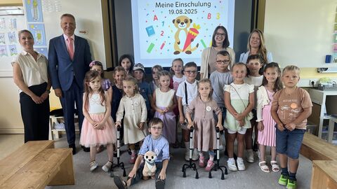 Mehrere Schülerinnen und Schüler stehen gemeinsam mit Erwachsenen in Klassenzimmer