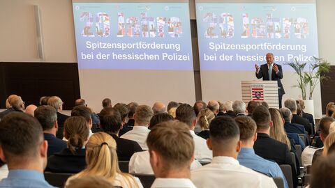 Eine Person steht auf der Bühne und hält eine Rede. Davor befindet sich Publikum.