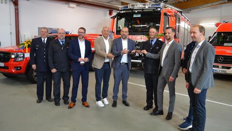 Neun Männer stehen in einer Reihe in einer Fahrzeughalle - im Hintergrund stehen drei Feuerwehrfahrzeuge.