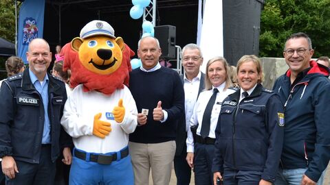 Minister Poseck steht mit fünf Polizeibeamten sowie dem Polizeimaskottchen in einer Reihe - im Hintergrund sieht man eine Bühne
