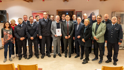 Heimatschutzminister Roman Poseck nach der Bescheidübergabe u. a. mit Bürgermeister Thomas Ciesielski Über 20 Männer und Frauen stehen teilweise in Anzügen und teilweise in Feuerwehrkleidung nebeneinander. Ein Mann in der Mitte hält den Zuwendungsbescheid hoch.