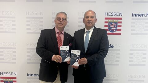 Innenminister Roman Poseck mit dem Vorstandsvorsitzenden der Jüdischen Gemeinde Gießen Lawrence de Donges-Amiss-Amiss Zwei Männer stehen nebeneinander und halten jeweils einen Flyer in den Händen.
