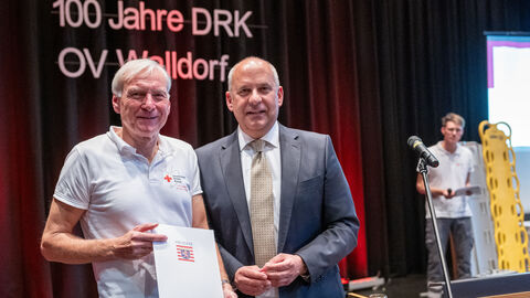 Zwei Männer stehen nebeneinander, einer hält einen Förderbescheid in den Händen. Im Hintergrund der Schriftzug "100 Jahre DRK OV Walldorf"