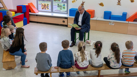 Roman Poseck sitzt auf einem Stuhl und liest ein Buch vor. Vor ihm sitzen Kinder auf zwei Turnbänken und schauen zu ihm.