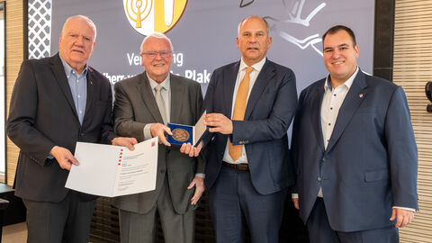 Vier Männer in Anzug stehen nebeneinander und halten Urkunde und Plakette in der Hand