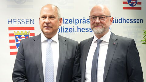 Zwei Männer im Anzug stehen vor Hessenwappen.