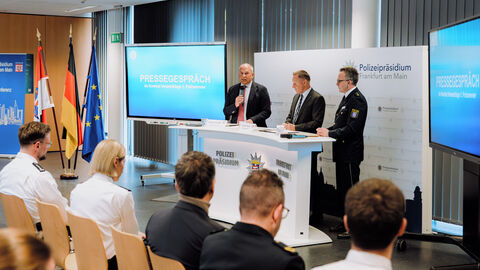 Drei Männer stehen an einem Rednerpult - Darauf bedruckt steht "Polizeipräsidium Frankfurt am Main"