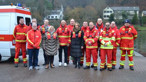 Eine Gruppe von Einsatzkräften der DLRG steht gemeinsam mit Roman Poseck und weiteren Zivilisten vor einem Einsatzfahrzeug am Ufer eines Flusses. Die Einsatzkräfte tragen rote Schutzkleidung mit gelben Streifen.