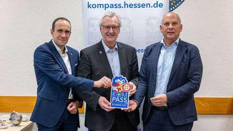 Drei Männer in Anzügen stehen nebeneinander. Der mittlere Hält ein Schild mit der Aufschrift "KOMPASS" in den Händen.