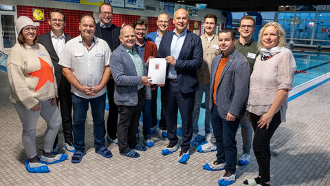 Innenminister Poseck hält einen Zuwendungsbescheid in den Händen und steht mit einer Gruppe von circa 10 weiteren Personen in einem Hallenbad.