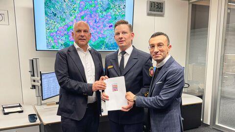 Drei Männer stehen nebeneinander in einem Büro und halten einen Förderbescheid. Hinter ihnen zeigt ein großer Bildschirm eine Landkarte mit markierten Standorten von Sirenen.