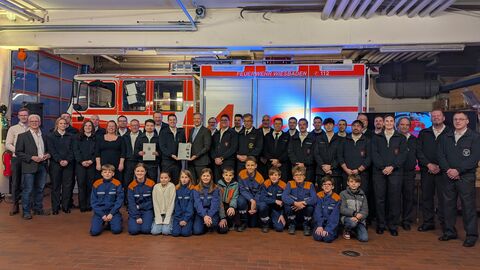 Mehr als 20 Personen stehen nebeneinander in ihrer Feuerwehrkleidung vor einem Feuerwehrauto und halten eine Urkunde hoch. Davor sitzen circa zehn Kinder, ebenfalls in ihrer Feuerwehrkleidung.
