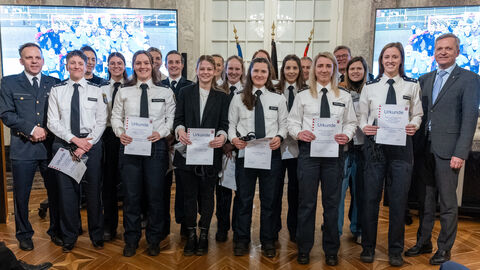 Gruppe von Polizistinnen in Uniform steht in einem festlichen Saal und halten Urkunden in den Händen. Sie posieren gemeinsam mit zwei weiteren Beteiligten vor Fahnen und Bildschirmen während einer Auszeichnungsveranstaltung.