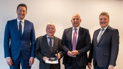 Vier Männer in Anzug nebeneinander vor einer weißen Wand - ein Herr hält eine Plakette in den Händen