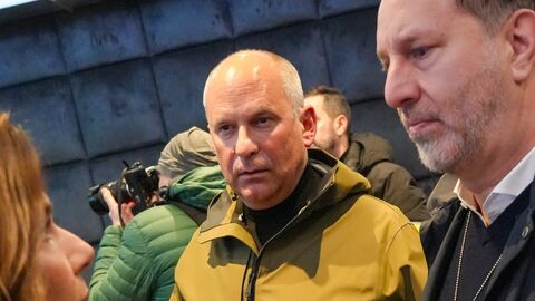 Zwei Männer sprechen am Abend im Freien mit einer Frau. Einer trägt eine gelb-schwarze Jacke, der andere einen Dienstausweis. Im Hintergrund steht ein Fotograf mit Kamera.