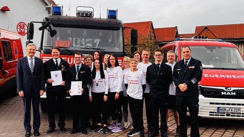 Mehrere Mitglieder der Feuerwehr sowie Innenstaatssekretär Martin Rößler stehen vor einem Feuerwehrauto und halten eine Urkunde hoch.