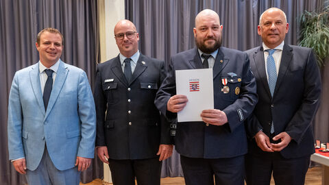 Vier Männer stehen bei einer Feierstunde nebeneinander auf einer Bühne. Ein Feuerwehrmann in Uniform hält eine Urkunde mit dem Wappen von Hessen in den Händen, neben ihm stehen weitere Vertreter der Feuerwehr sowie Innenminister Roman Poseck.