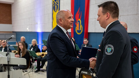 Roman Poseck reicht Wehrführer Jan Kettler auf einer Bühne die Hand. Im Hintergrund sieht man die Big Band der Feuerwehr. 