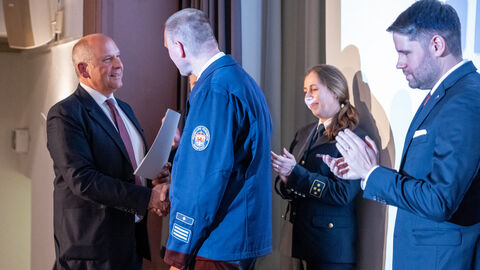 Roman Poseck überreicht Wehrführer Marcus Sattler eine Auszeichnung, Feuerwehrangehörige klatschen im Hintergrund.