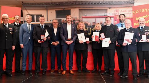 Mehrere Feuerwehrangehörige in Uniform und Gäste aus Politik und Verwaltung stehen auf einer Bühne mit rotem Hintergrund. Einige halten Urkunden und Auszeichnungen in den Händen. Im Hintergrund ist ein Banner zu „50 Jahre Jugendfeuerwehr“ zu sehen.