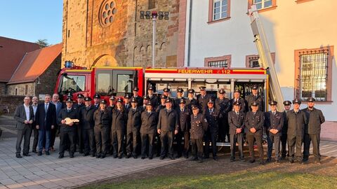 Gruppenfoto von Feuerwehrangehörigen und Vertretern aus Politik und Verwaltung vor einem roten Löschfahrzeug mit ausgefahrener Leiter. Die Einsatzkräfte tragen Uniform, im Hintergrund sind historische Gebäude zu sehen.
