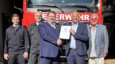 Fünf Männer stehen lächelnd vor einem großen, roten Feuerwehrfahrzeug in einer Fahrzeughalle, während die beiden mittleren Männer ein offizielles Dokument halten und sich die Hand schütteln.
