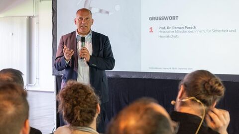 Herr Poseck steht vor einer Leinwand, auf der eine Powerpoint Präsentation zu sehen ist, und spricht ins Mikrofon.