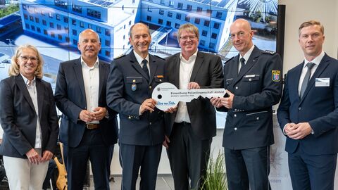 Innenminister Poseck steht gemeinsam mit fünf weiteren Personen in einer Reihe - zwei von ihnen tragen schicke Polizeiuniformen - und drei der Personen halten gemeinsam einen überdimensionalen Schlüssel in den Händen