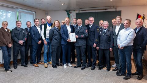 Herr Poseck steht mit einer Gruppe von knapp 20 Personen in einem Raum - gemeinsam mit einem Mann in eleganter Feuerwehruniform hält er einen Bescheid in den Händen.