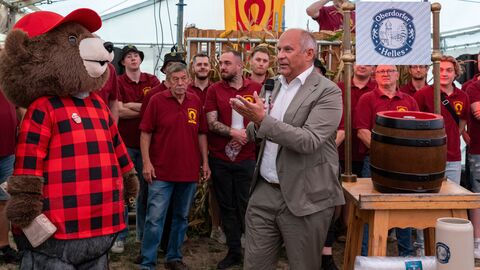 Herr Poseck hält eine Rede - im Hintergrund eine Gruppe von Männern mit weinroten T-Shirts, neben ihm ein Fass Helles und auf der anderen Seite eine Person in einem Bären-Maskottchen-Kostüm mit einem Hammer in der Hand um das Fass anzustoßen