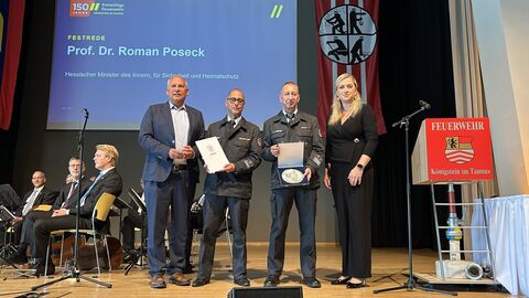 Herr Poseck steht mit zwei Herren in festlicher Polizeiuniform und einer Dame auf einer Bühne - neben ihnen sieht man ein Orchester und im Hintergrund einer Powerpoint mit der Aufschrift "Festrede - Prof. Dr. Roman Poseck"