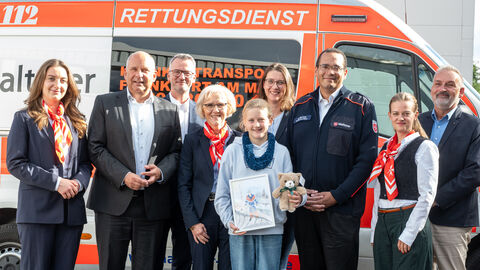 Herr Poseck steht mit 7 Erwachsenen und einem Mädchen vor einem Rettungswagen der Malteser. Das Mädchen hält ein selbstgemaltes Bild in den Händen.